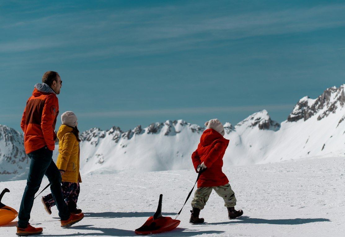 Família com crianças puxando trenós na neve, montanhas ao fundo
